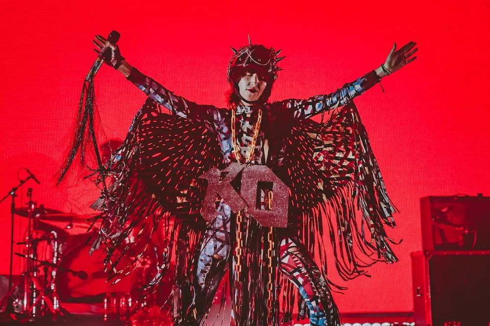Karen O with a spiky helmet, fringed cape, huge KO letters on her front performing with her arms stretched out