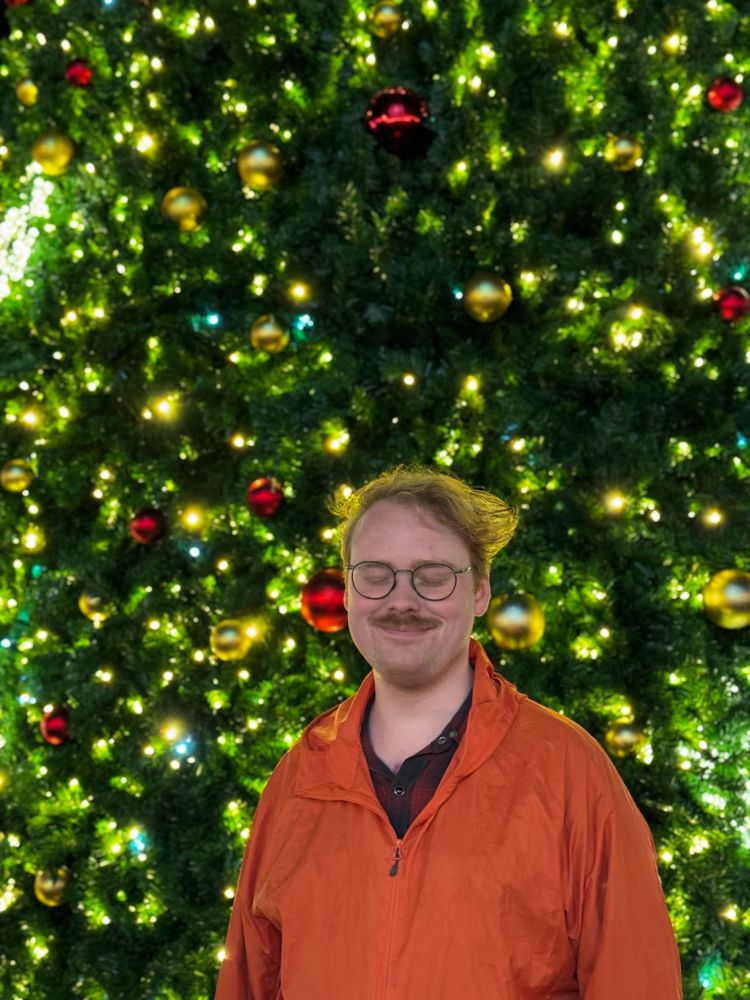 Mischa standing in front of a large Christmas trees with their eyes closed 