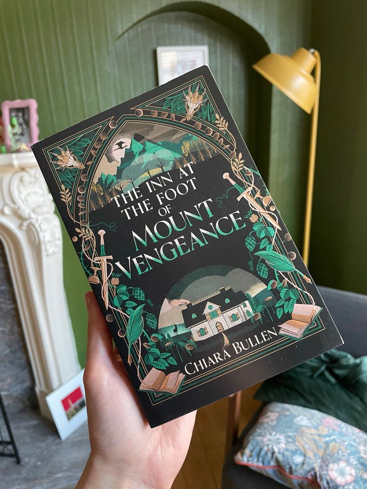 The UK edition of my novel The Inn At The Foot Of Mount Vengeance, held up against my living room. There is a green wall and a yellow lamp behind it, as well as a white fireplace. The book cover is mostly black, with the title in the middle. Above is the image of a green mountain. Below there is a white inn. The edges are framed by an array of ivy, thistles, daggers, quills and books. 