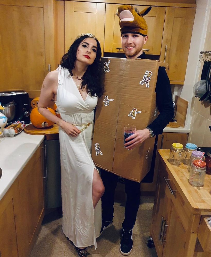 A woman with brown curly hair in a crushed, cheap white dress and a golden tiara. Her boyfriend wears a literal cardboard box with ‘Greek soldier’ stickers on it. He is wearing a horse hat. 