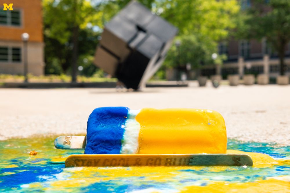 A maize and blue popsicle melting and a popsicle stick that reads “Stay Cool and Go Blue” with a Block M in front of The Cube