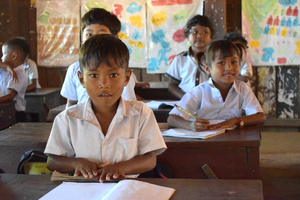 A mid close shot of a school boy sitting at a classroom desk with text book on the table. Four more students sit behind him. At a rural Ponheary Ly Foundation primary school in northern Cambodia