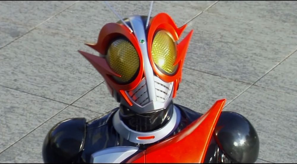 a photo of kamen rider g from the shoulders up. he has black shoulders with a small silver and large red pair of triangle shapes on his chest. his helmet has a sliver face plate with large translucent yellow bug like eyes with sharp red g's around them and a pair of silver antenna.