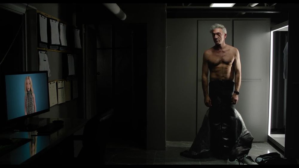 a screenshot from the 2024 david cronenberg film "the shrouds", with the main character karsh (player by vincent kassel) standing at the right edge of the screen standing shirtless wearing one of the large black leather titular shrouds. at the left side of the screen, is a cgi blonde woman on a computer screen.