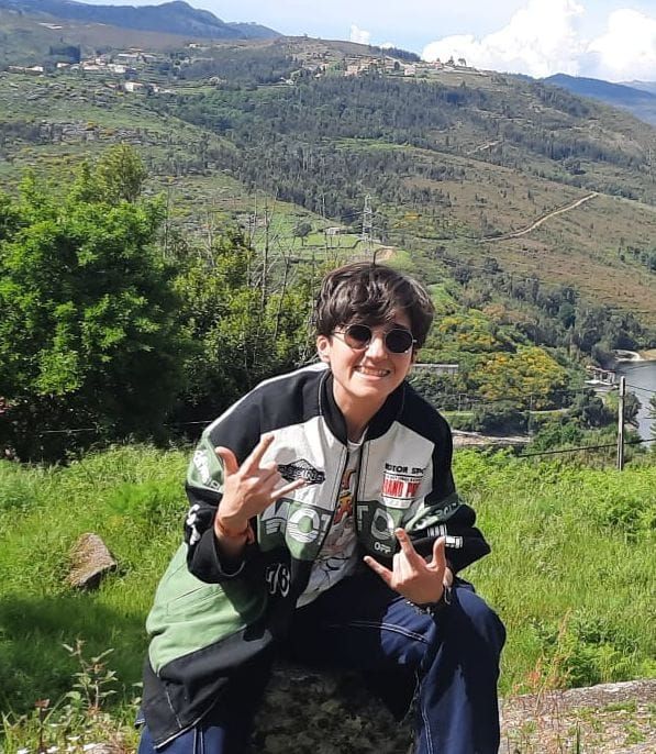 Person with brown short hair, wearing sunglasses, a green racing jacket, a white shirt, and jeans sitting on a rock. They're smiling and doing this gesture 🤘🤘
They're somewhere in the country side, judging by the green fields and mountains on the background.