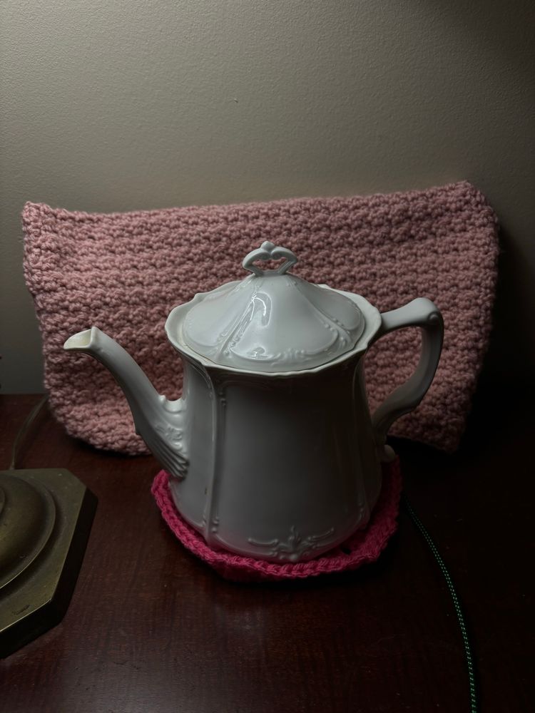 A white china teapot on a dark rose yarn trivet with a lighter pink crocheted envelope behind it. The base of a brass lamp is on the left. 