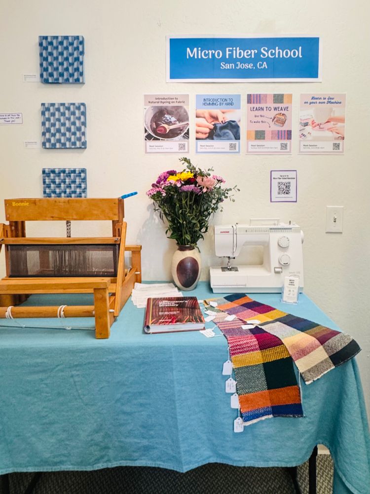 Several signs describing upcoming classes hang on the wall in front of a table with a loom, sewing machine, and samples.