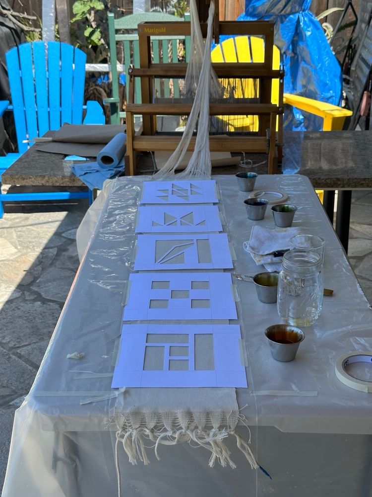A woven cloth is laid on a table covered with plastic sheeting. It's connected to a table loom named "Marigold" on one slide. There are stencils with geometric shapes placed on top and small jars of paint are scattered on the table.