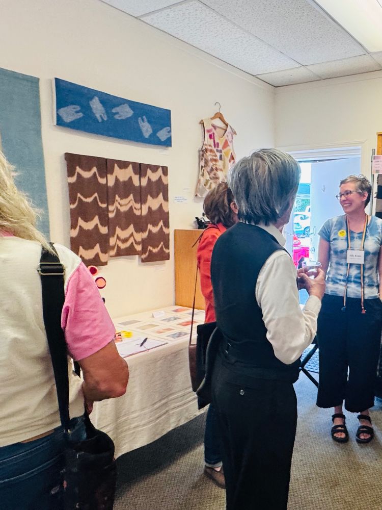Ally standing talking to several visitors in her studio. There are two ikat wall hangings and a painted warp top hanging on the wall.
