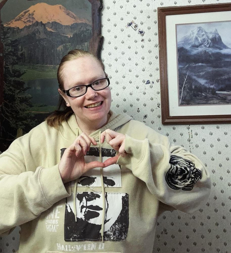 Photo of me, smiling and doing heart hands, I’m wearing my Michael Myers hoodie and have my hair in a ponytail