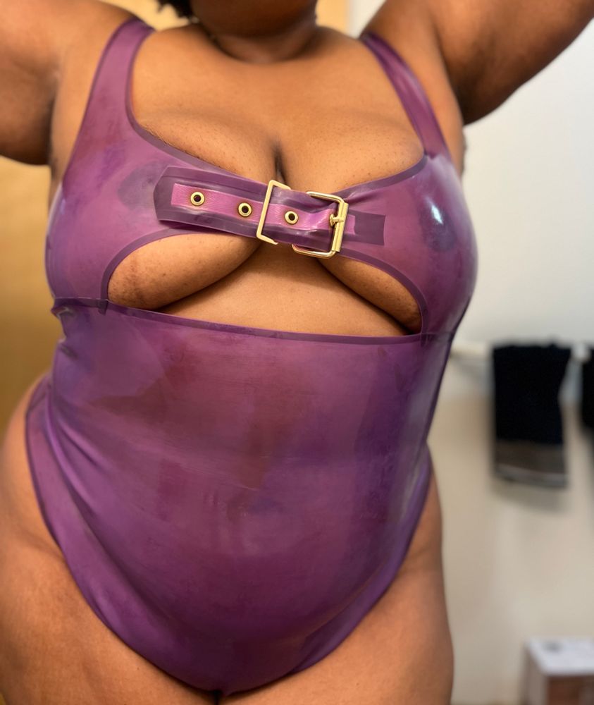 A close up of Dolly’s ample curves poured into a translucent purple latex bodysuit. There is a buckle across the chest holding her perfect tits high and tight.