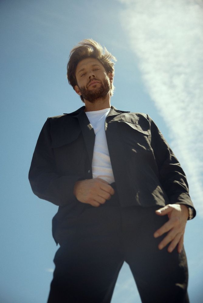 Jensen Ackles in black suit and white shirt with his hand on the waist of his pants against blue sky 