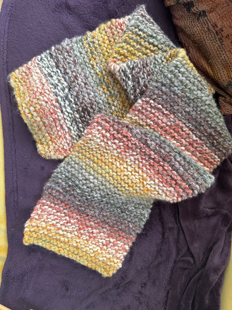 Loosely folded, bulky handknit, multicolored scarf, subtly striped in shades of mustard yellow, brown, rust, olive green, and cream. Sitting on eggplant purple blanket background.
