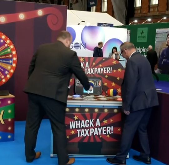 Two middle aged men in suits engage in a fairground type game at Conservative Party Conference titled 'WHACK A TAXPAYER!' An attempt to make some sort of point about Labour spending plans. Probably.