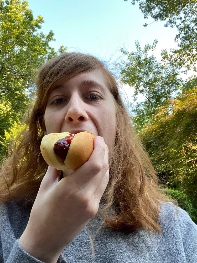 A selfie with the subject devouring a weiner (hot dog)

The individual has red shoulder length hair and is standing outside amongst a bunch of trees 