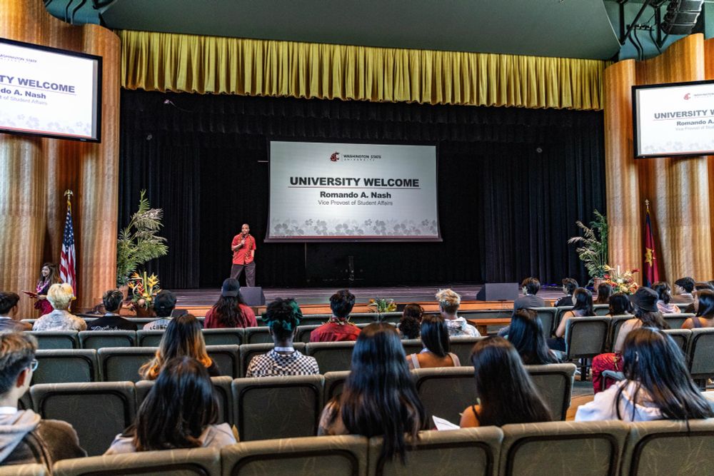 Color indoor photo of a presenter on a stage with people in an auditorium watching 