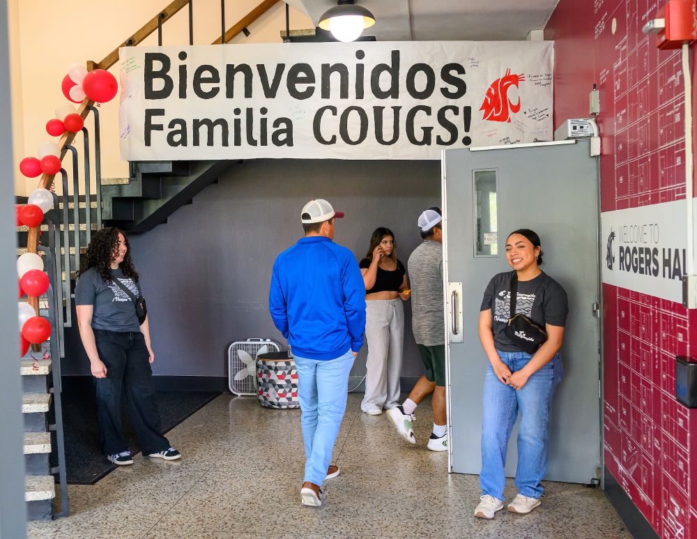 Color indoor photo showing people standing in front of a door and people going through the door.  A large sign above reads "Bienvenido's Familia Cougs!" 