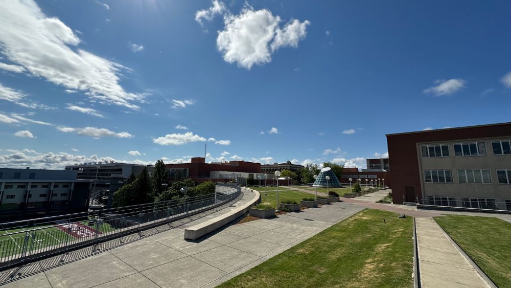 Color outdoor photo showing green grass and several academic buildings. Blue sky and white clouds are above. 