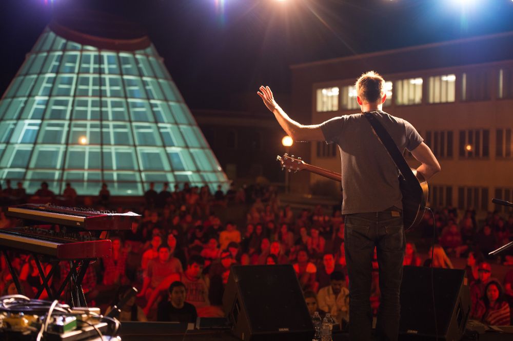 Color outdoor photo of a performer on a stage with a guitar with his back to the camera. A large crowd is in front with a blue/green glass atrium lit up in the background. 