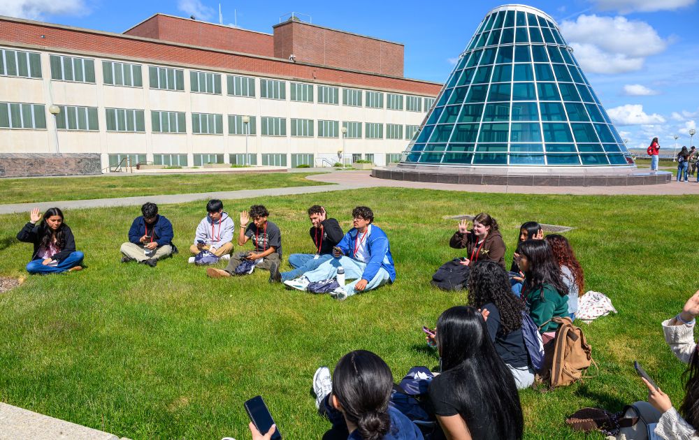 Color outdoor photo of a group of students. They are sitting on green grass with various people and buildings in the background. 