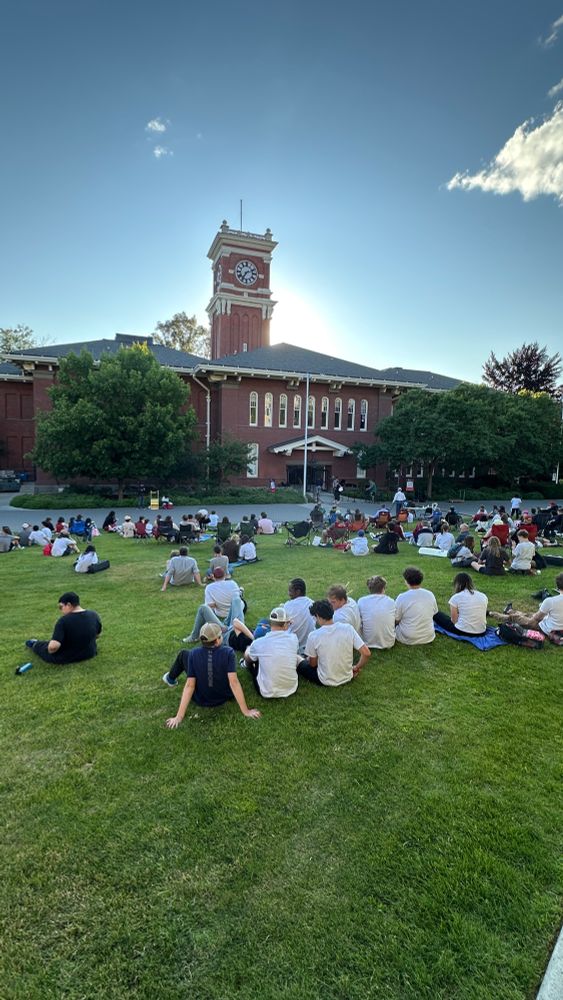 Color outdoor photo during daytime showing people sitting on a green grass field watching a concert in front of them