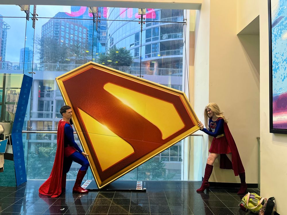 A Superman cosplayer and a Supergirl cosplayer pushing the red and yellow logo from the new movie from opposite sides of it. The Superman cosplayer is in a blue suit with red boots, trunks, and a cape. The Supergirl cosplayer has a blue long-sleeved bodice with a red and gold logo, red skirt, red boots, and a red cape. 