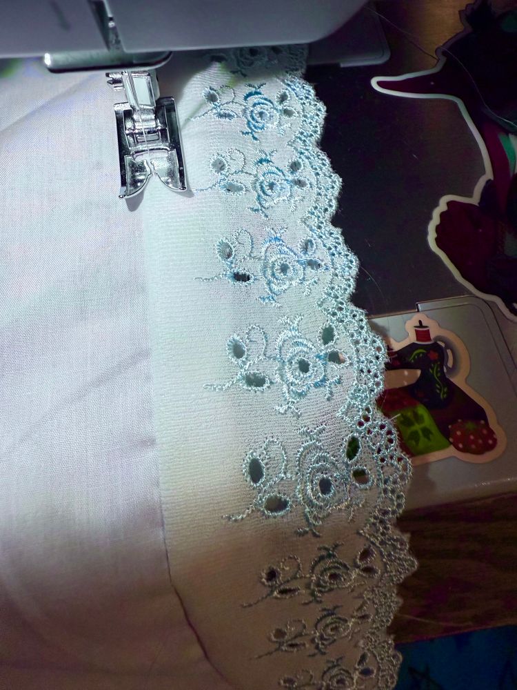 White and blue eyelet lace trim being stitched onto some white polyester broadcloth via sewing machine. The eyelet lace is white with blue thread stitched into rose patterns.