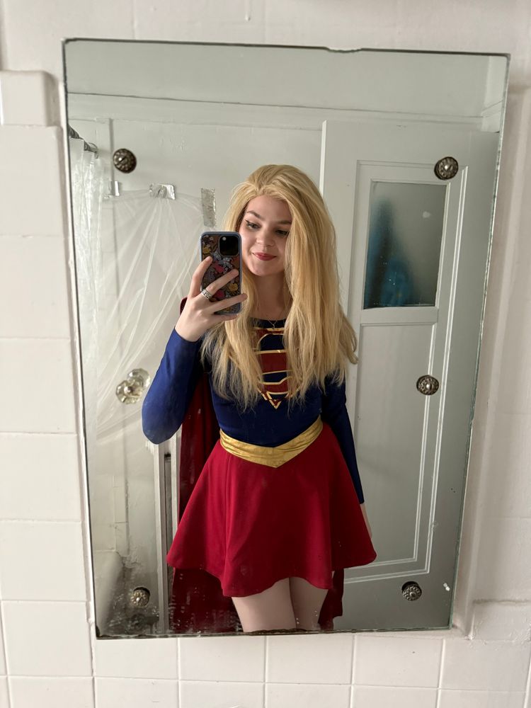 Another mirror selfie of me in my Supergirl cosplay with the blonde wig, dark blue bodice, gold belt, red miniskirt, and red cape, and have done my makeup. 

In the background there is a plastic tarp taped to my wall for very long and complicated reasons that shorten to: My Landlord Sucks

And I'm holding an entirely different phone because yesterday I wound up upgrading mine and getting thoroughly roasted for being a Nerd the entire time by the employee helping me transfer my data. This phone has a pink and blue floral otterbox case