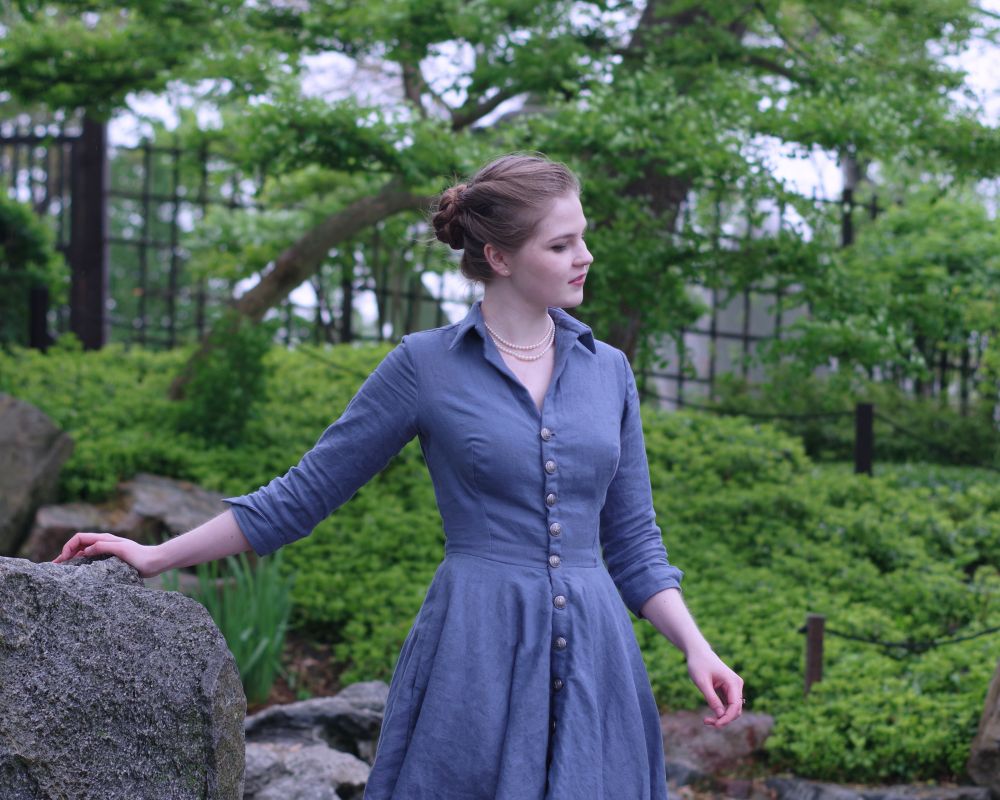 A photo of me standing in a garden with my hand on a rock. I'm wearing a steeley blue linen dress, with 3/4 length sleeves, a collar, and buttons up the front. It has about a knee-length circle skirt and a very vintage feel to it. My hair is also styled up in a braided bun.