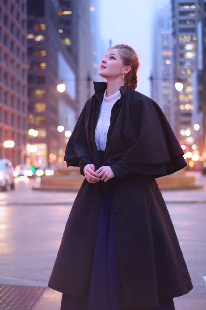 A photo of someone standing in the street in Chicago looking up at the buildings. They're wearing a long dark gray coat with two laters of elbow-length capes atop them. They're also wearing a long navy skirt and a white blouse.