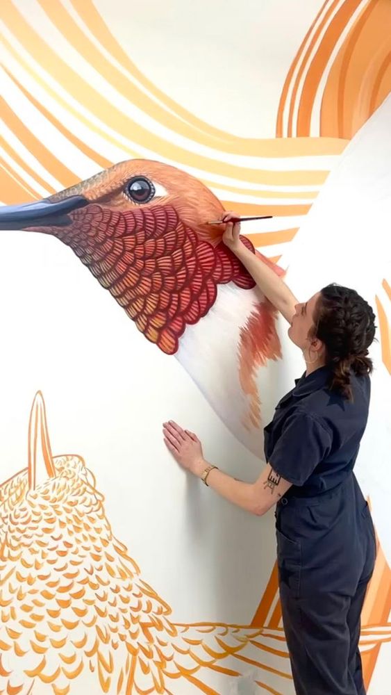 I am wearing a grey jumpsuit and am standing on my tiptoes to paint the hummingbird’s gorget (colorful throat feathers). 