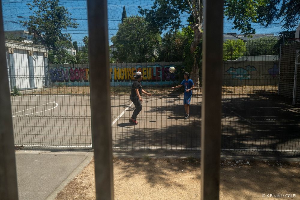 Deux adolescents jouent au foot dans la cour grillagée d’un centre éducatif fermé. Ils jouent sur un terrain de foot, qui peut aussi servir de terrain de basketball. Des fresques sont peintes sur le mur en arrière plan. 