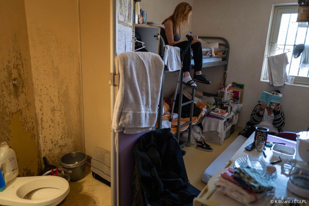 Cellule surpeuplée d’un centre pénitentiaire dans laquelle trois femmes sont présentes. Une est assise sur un matelas par terre en train de lire un livre, deux autres sont dans leur lit superposé. Une est assise et est en train d’écrire, une béquille est suspendue à côté d’elle. La dernière est allongée dans son lit. On peut voir leur sanitaire qui est insalubre, avec des traces de moisissure. 