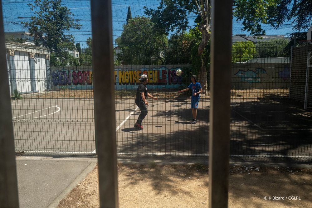 Deux adolescents jouent au foot dans la cour grillagée d’un centre éducatif fermé (CEF). Ils jouent sur un terrain de foot, qui peut aussi servir de terrain de basketball. Des fresques sont peintes sur le mur en arrière plan. 