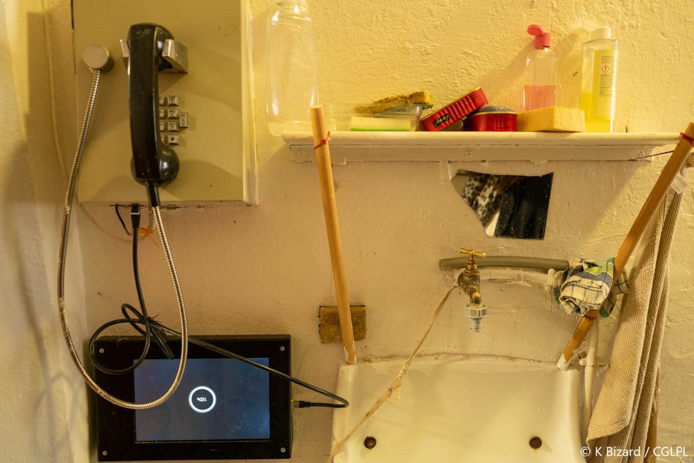 Point phone installé dans une cellule. Le téléphone mural se trouve à gauche de la photo, une tablette de visiophonie est également fixée au mur sous le téléphone. Un lavabo et une étagère se trouvent juste à côté du téléphone, à la droite de la photo