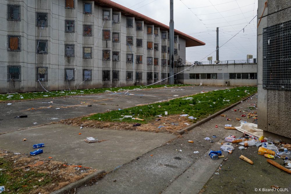 Photo de l’extérieur d’un centre pénitentiaire, le sol est rempli de déchets (pain, sacs plastiques, briques de lait…). Dans le fond de la photo, on voit la façade du centre pénitentiaire.