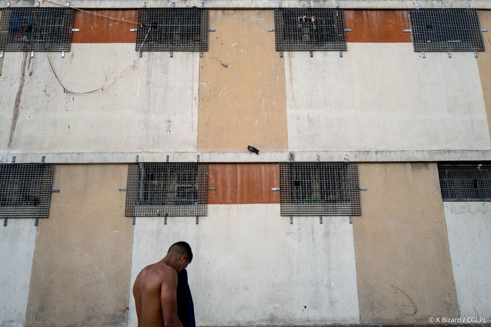 Vue depuis la cour de promenade d’une maison d’arrêt sur le mur des cellules devant lequel se trouve un homme de dos, torse nus. Les fenêtres des cellules sont équipées de caillebotis (grillages en fer).