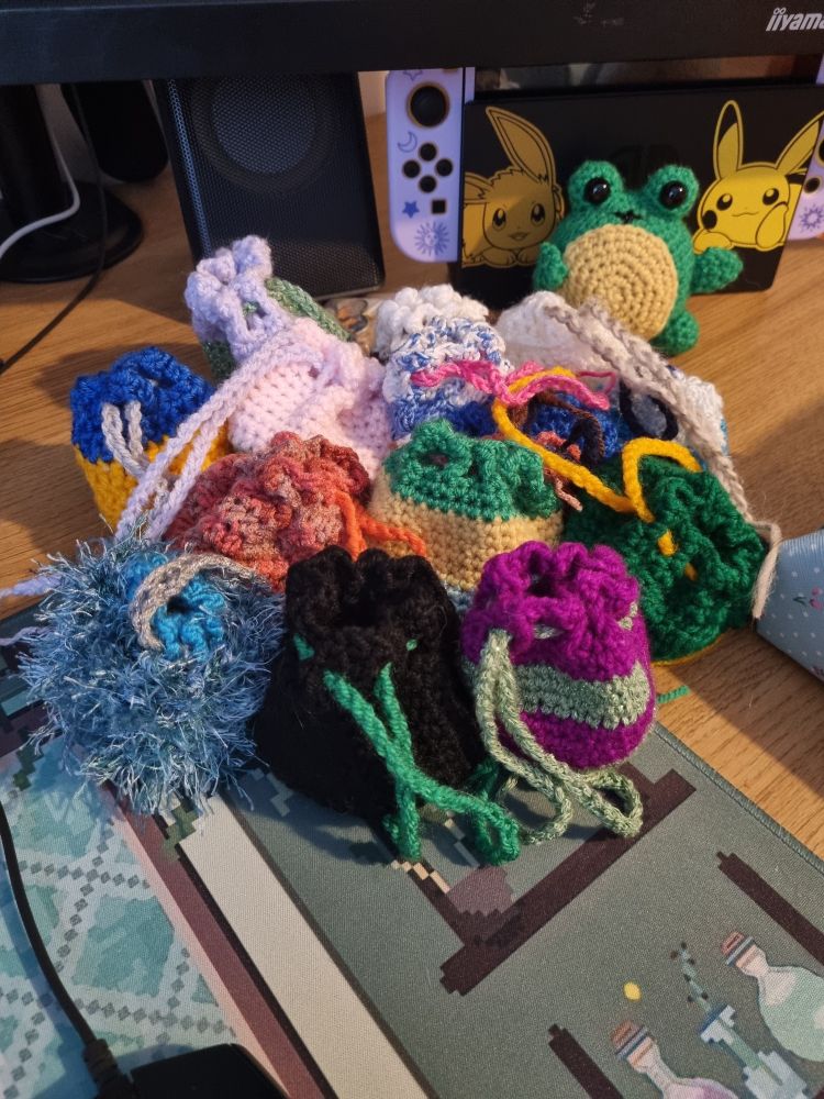 various dice bags made by hand with crochet and various fibers of different colours. Most are striped in dual colours that match the dice in their bag. There's also a crochet frog waving from the background. Cute.