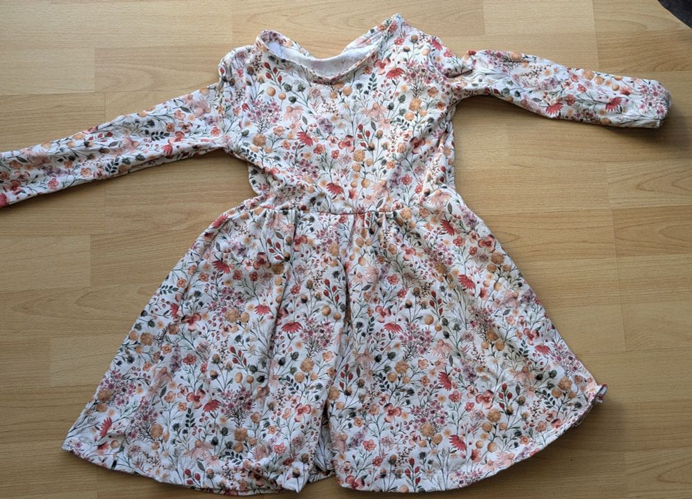 Auf einem hellen Holzfußboden liegt ein Kinderkleid mit hellem Stoff, auf dem viele kleine Blumen gedruckt sind