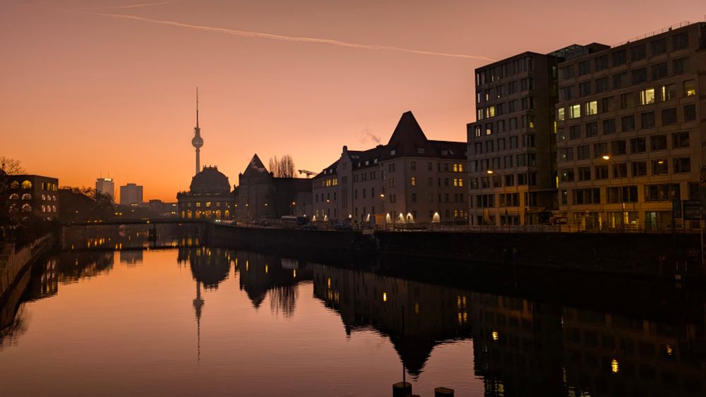Ein Blick im Sonnenaufgangslicht über die Spree. Im Hintergrund sind das Bode-Museum sowie der Fernsehturm zu sehen. Rechts im Bild stehen Bürogebäude.
Alles spiegelt sich im unbewegten Wasser des Flusses.
