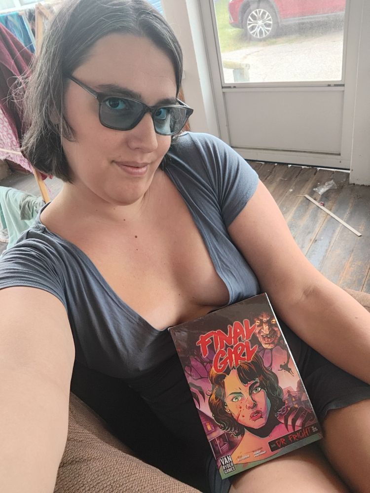 Yvie is sitting on a couch on the porch. She is wearing a dark gray blue dress that shows off her boobs as well as a pair of clear gray glasses with blue lenses. She has a copy of the game final girl on her lap. It is Frightmare on Maple Lane scenario 