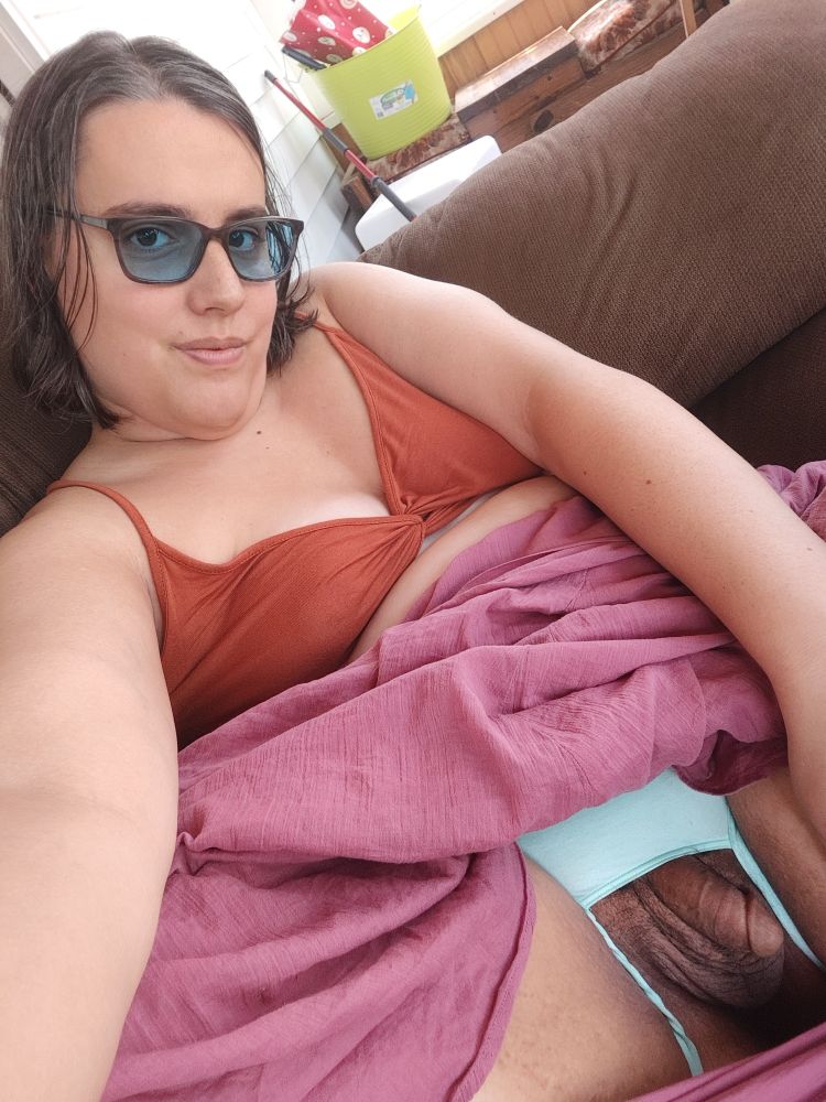 Yvie is laying on a couch.  She is wearing an orange crop top and a pink skirt that she has pulled up to show her light blue green bottomless panties and her girl cock. 