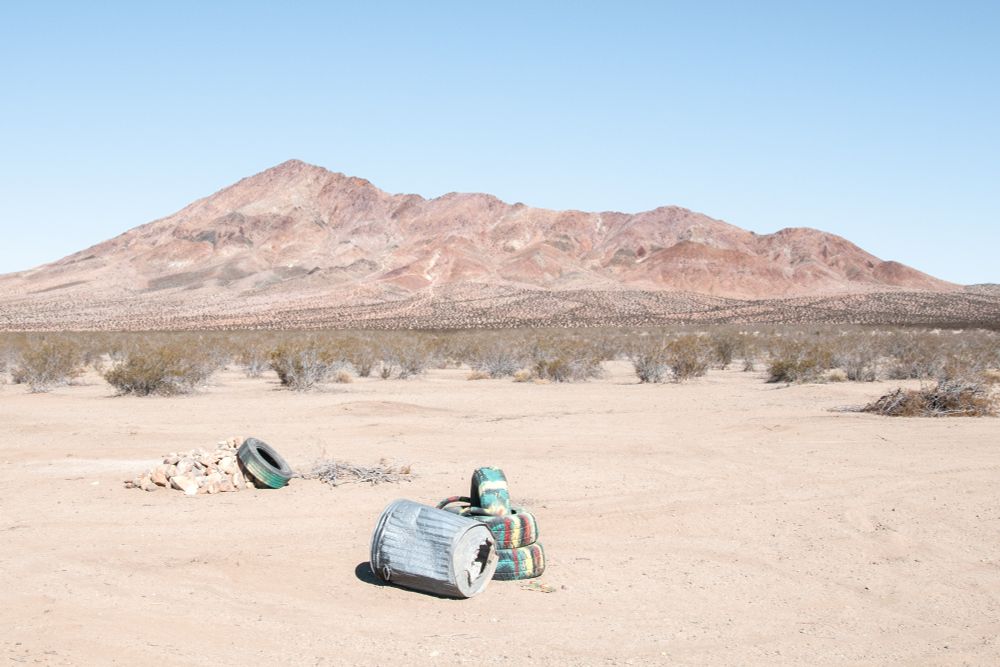 A reddish desert peak stands alone behind a scrubby desert plain. On that plain in a clearing in the foreground, some painted tires and a garbage bin.