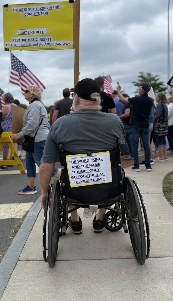 a sign on the back of a protester’s wheel chair that says “the word king and the name trump only go together as ‘fucking trump’”