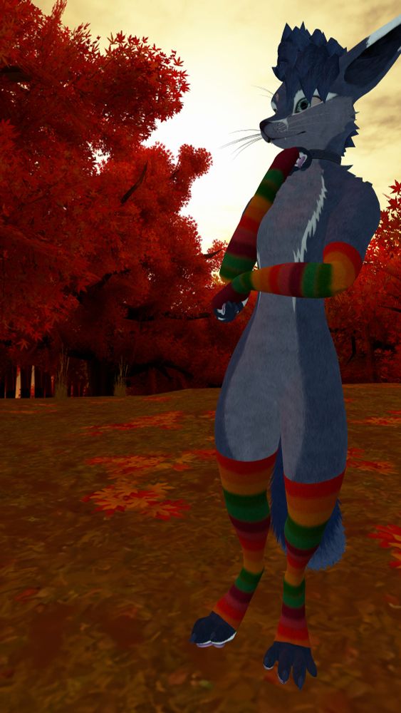 In-game photo of a fox-like anthropomorphic VRChat avatar with cyan eyes, dark blue hair, blue fur mixed with light blue markings around the eyes, on the muzzle and across the chest, and pink paw pads. The character is wearing a dark blue collar and rainbow-striped arm and leg warmers. They are looking off to their right side with one arm supporting the other, while their fist is pressed against their chin, as if lost deeply in their thoughts. The background contains an autumn scene with trees whose leaves are orange, with some leaves scattered around the ground, set in front of a bright yellow sky.