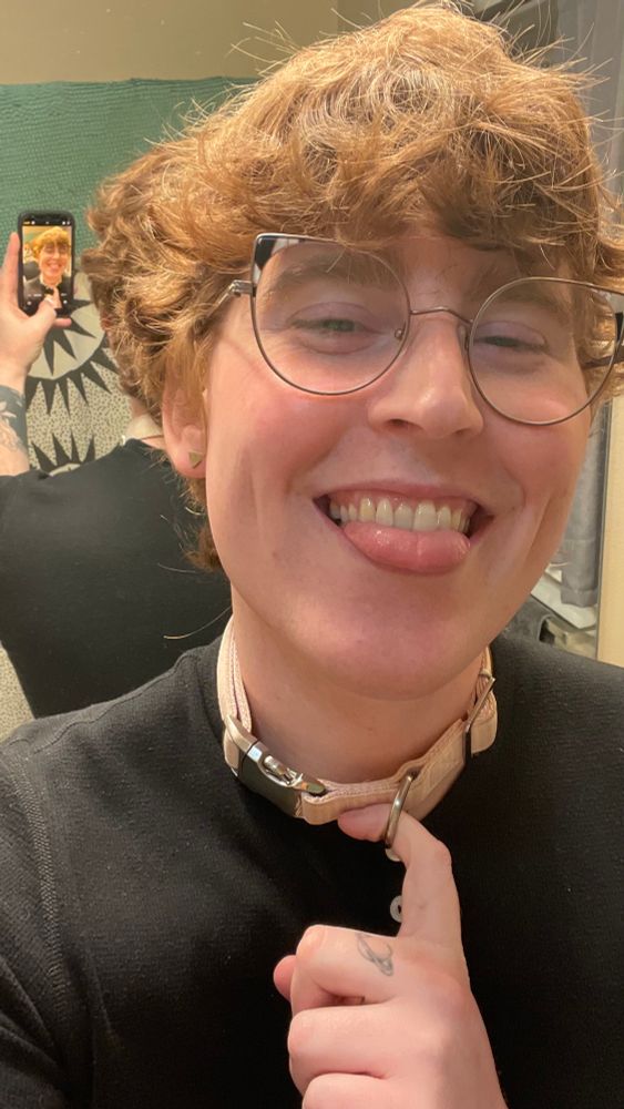 Kezia, smiling with her tongue out, wearing a pink collar with one index finger hooked in the D ring. 