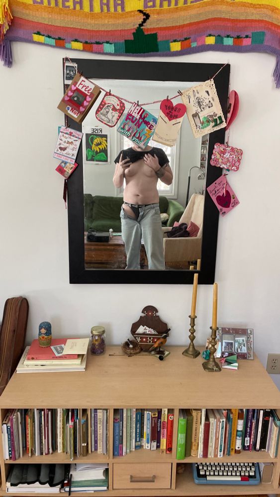 Mirror selfie of a half undressed Kezia. The mirror is festooned with handmade cards congratulating a friend on top surgery. On card obscures faer face. 