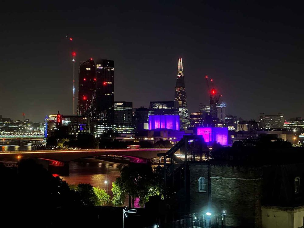 A photograph of the London skyline from around Trafalgar Square. In the photograph the Themes is visible in the foreground with  the National Theatre behind illuminated in purple. There are numerous high rise buildings in the background including the Shard with various office lights on. Red anti aircraft lights are illuminated. 