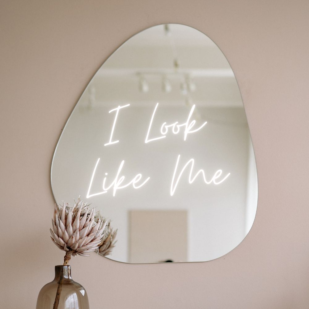 A flower sits in a vase in front of a mirror against a cream-colored wall. The words “I Look Like Me” and illuminated in the reflection of the mirror. 