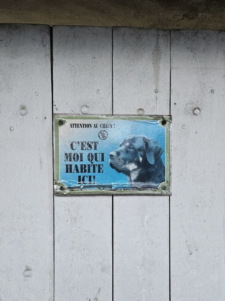 Placa presa em um portão de madeira clara mostrando a foto de um cachorro que parece ser da raça Rottweiler. A placa contém o aviso em francês 'Attention au chien! C’est moi qui habite ici!', indicando a presença de um cachorro na casa.